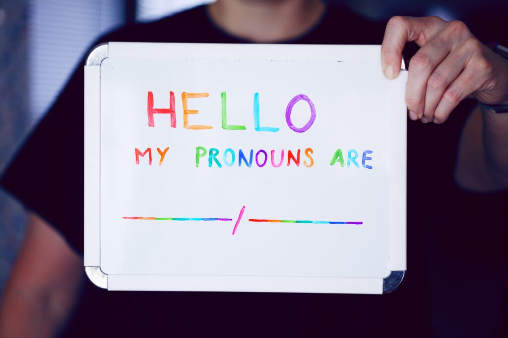 Person holding a small writing slate with Hello My Pronouns are in Pride colours