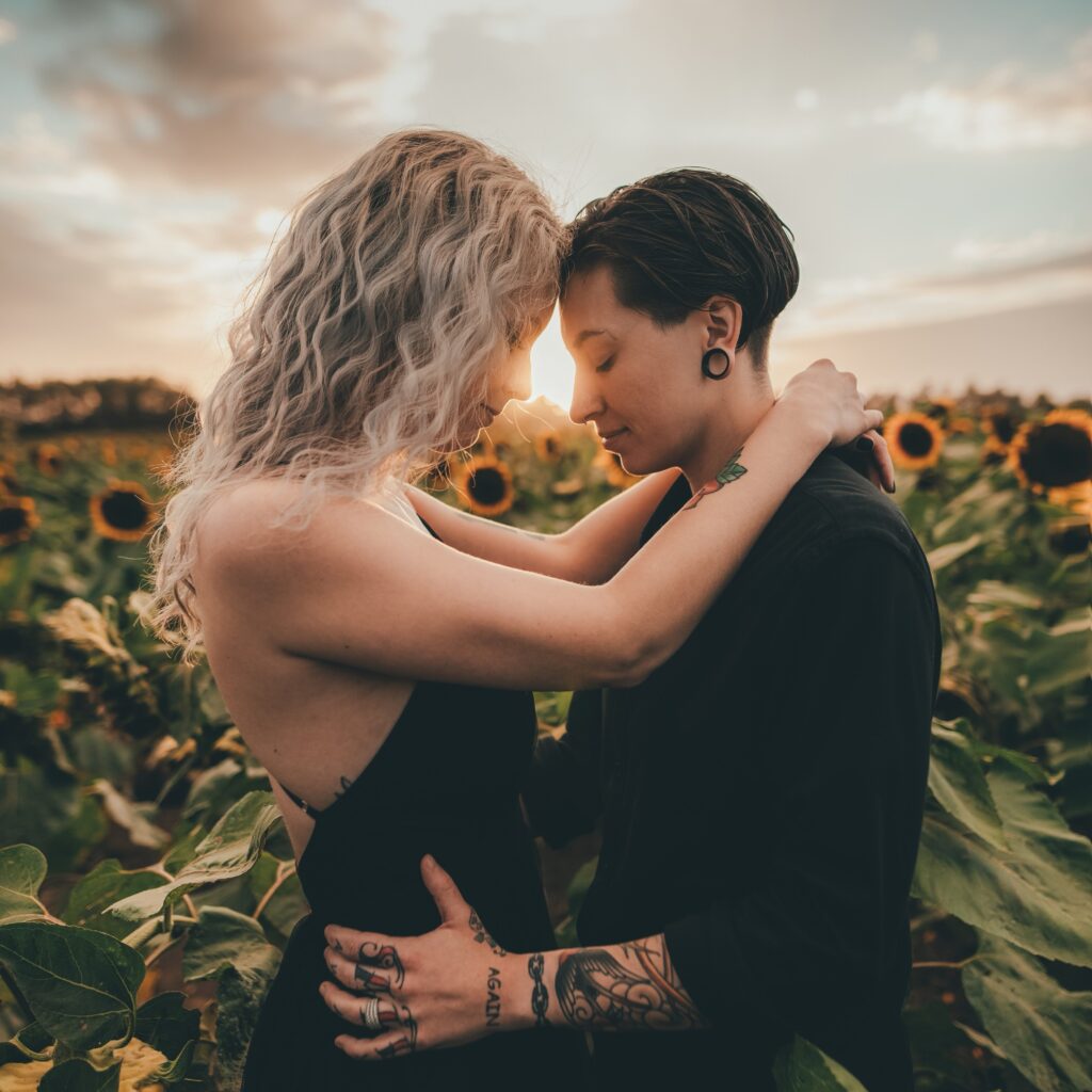 A non traditional couple standing in a field of sunflowers as the sun sets behind them.